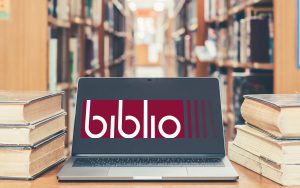 Notebook screen open in a library among old stacks of books. On the screen there is the biblio project logo